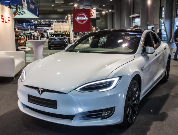 A white Tesla Model S on display at an automotive exhibition, featuring sleek aerodynamic lines, black alloy wheels, and a glossy finish. The car is showcased in a well-lit venue with other automotive brands, including Nissan, visible in the background.