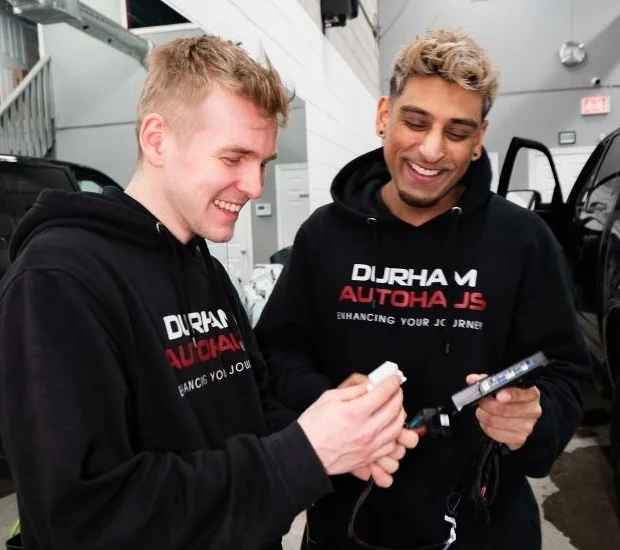 Two smiling men wearing black "Durham Autohaus" hoodies are inside a workshop, examining an electronic car component. One holds a device with a cable attached, while the other looks at it with interest. A black vehicle with an open door is visible in the background, along with white walls and workshop equipment.