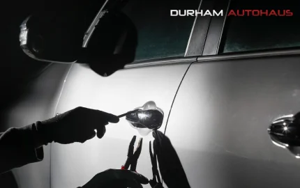 A shadowed figure wearing gloves uses tools to manipulate the lock of a silver car door at night. A flashlight illuminates the handle, casting dramatic shadows, while the Durham Autohaus logo appears in the top right corner. The image suggests a focus on car security, locksmith services, or theft prevention.