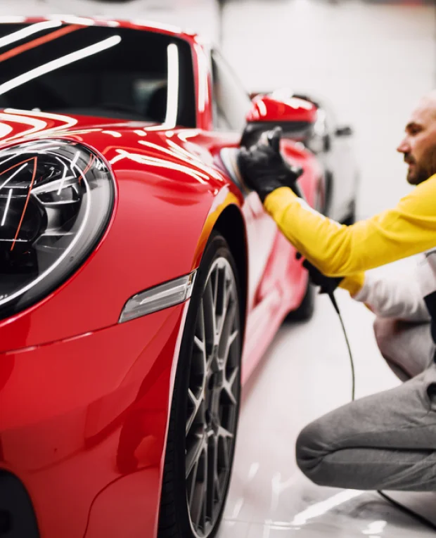 A professional detailer wearing black gloves and a yellow long-sleeve shirt uses a polishing machine on the sleek, red body of a luxury sports car. The car's glossy finish reflects the bright overhead lights in the detailing studio.