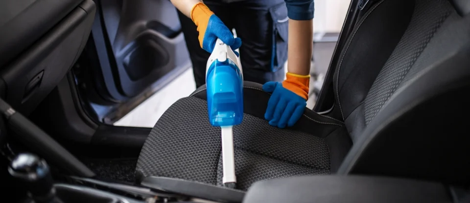 A professional cleaner wearing blue gloves and orange wrist cuffs uses a handheld vacuum to clean the fabric seat of a car. The car door is open, allowing light to highlight the detailing process.