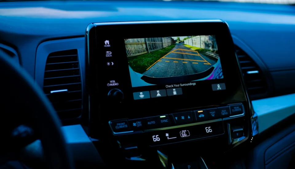 A car's infotainment screen displaying a rearview camera feed with parking guidelines and a warning to "Check Your Surroundings." The dashboard controls and air conditioning settings are visible below the screen.