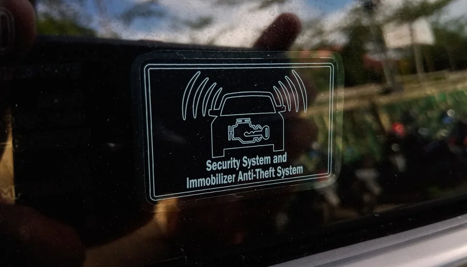 A close-up of a car window displaying a security system sticker that reads "Security System and Immobilizer Anti-Theft System." The sticker features an icon of a car with an engine symbol inside and wireless signal waves on both sides, indicating an active anti-theft system. A reflection of the photographer's hand and the surrounding outdoor environment is visible on the glass.