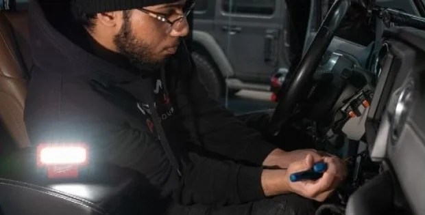 A man wearing a black hoodie, beanie, and glasses is seated in the driver's seat of a vehicle, using a tool to work on the ignition system. A bright portable light is illuminating the area, and another vehicle is visible in the background.