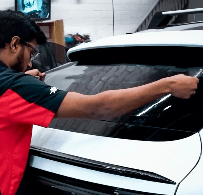 A technician wearing a black and red uniform carefully applies window tint film to the rear windshield of a white car. He stretches the film evenly across the glass, ensuring a smooth installation inside a workshop setting.