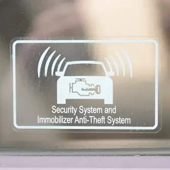 A car window sticker displaying "Security System and Immobilizer Anti-Theft System," with an icon of a car emitting signal waves and an engine symbol inside. The sticker indicates that the vehicle is equipped with an immobilizer to prevent unauthorized use.