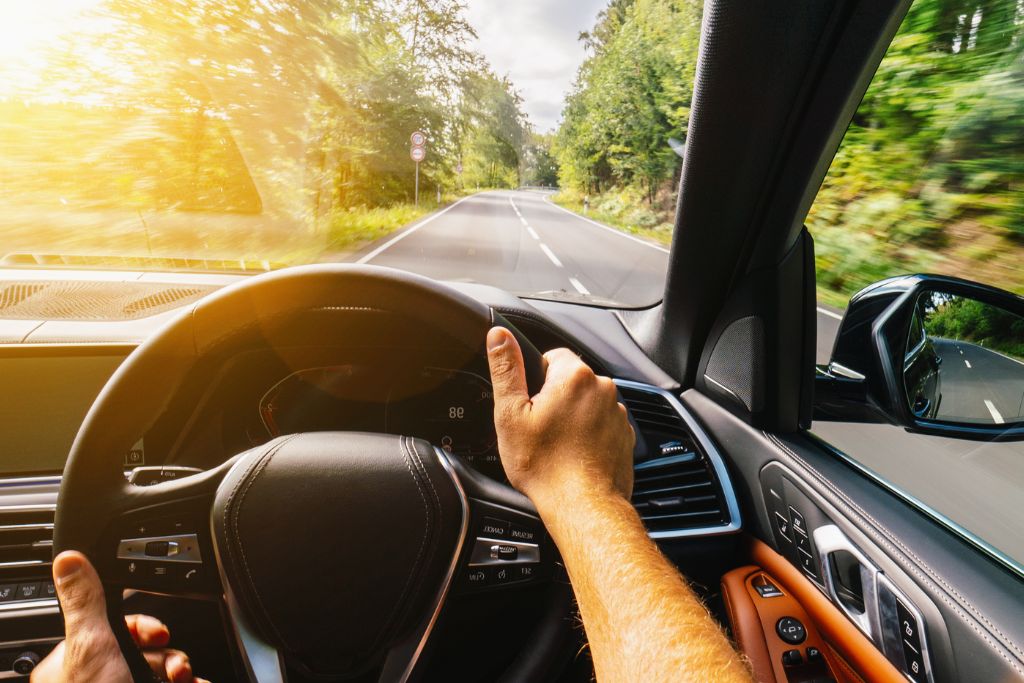A driver grips the steering wheel while bright sunlight streams through the windshield, illuminating the car's interior. The dashboard, side door, and road ahead are visible, with lush greenery lining the highway. The intense sunlight highlights the need for UV protection window film to reduce glare, prevent interior fading, and protect against harmful UV rays.