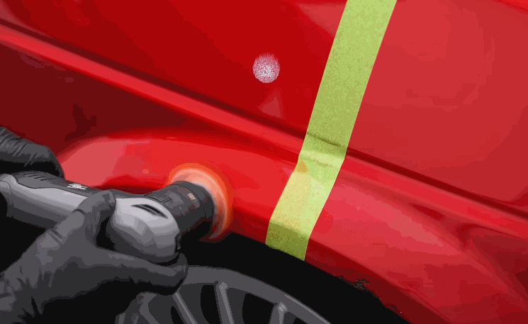 A detailer polishes the lower panel of a red car near the wheel well, with a green masking tape line marking the before-and-after section. The pad smooths out imperfections to restore clarity