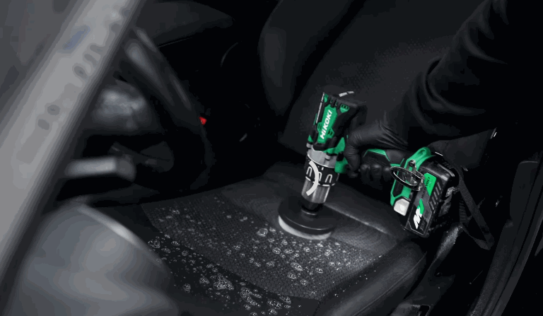A detailer uses a power drill with a brush attachment to deep-clean a foam-covered car seat. The interior is dark, and the process highlights precision upholstery care.