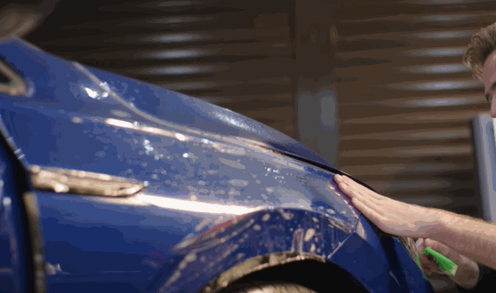 A technician presses down paint protection film over a wet blue car fender, ensuring proper adhesion.