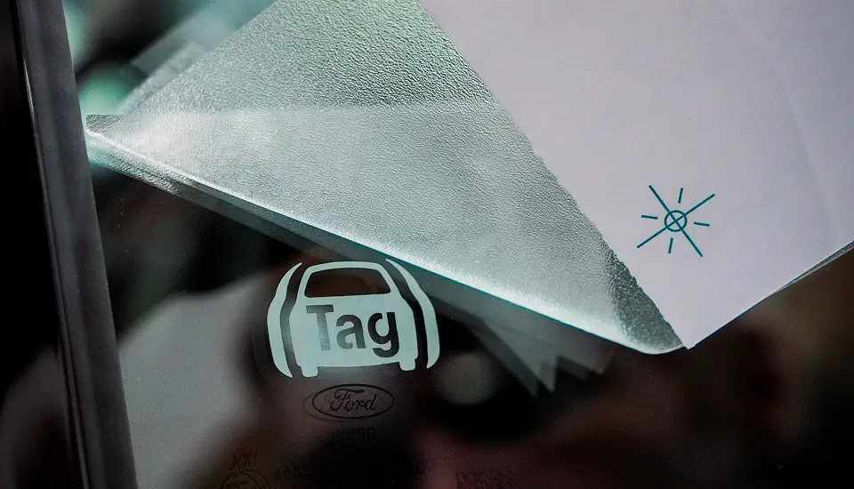 A close-up of a car window displaying a security system sticker that reads "Security System and Immobilizer Anti-Theft System." The sticker features an icon of a car with an engine symbol inside and wireless signal waves on both sides, indicating an active anti-theft system. A reflection of the photographer's hand and the surrounding outdoor environment is visible on the glass.